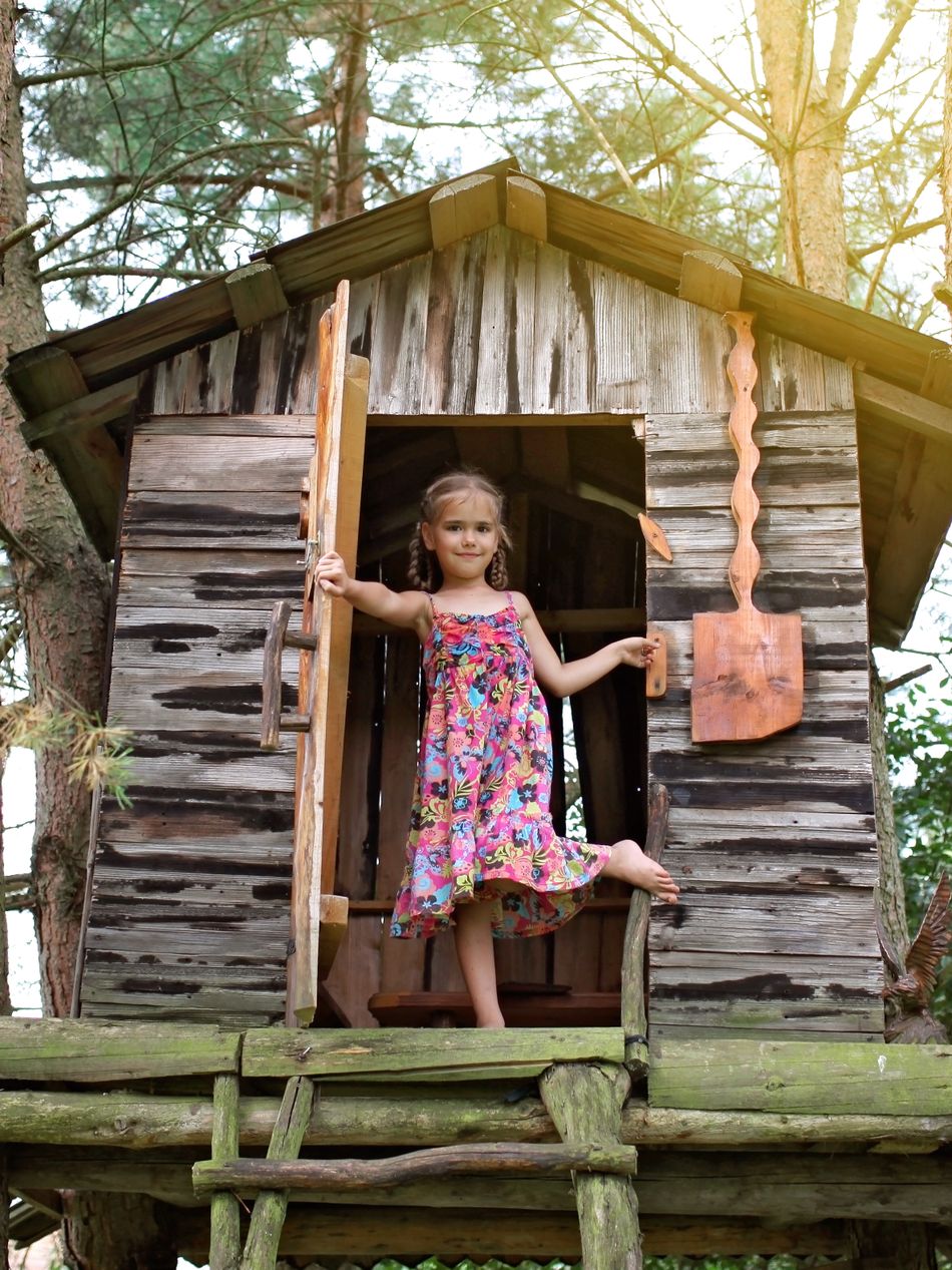 Mädchen mit zwei Zöpfen und einem geblümten Sommerkleid schaut aus der Tür eines Baumhauses.