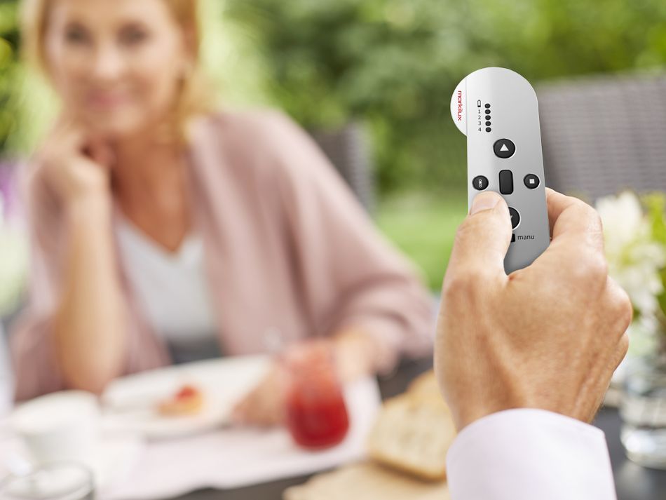 Mann hat markilux Fernbedienung an der Hand, Frau am Frühstückstisch im Hintergrund.